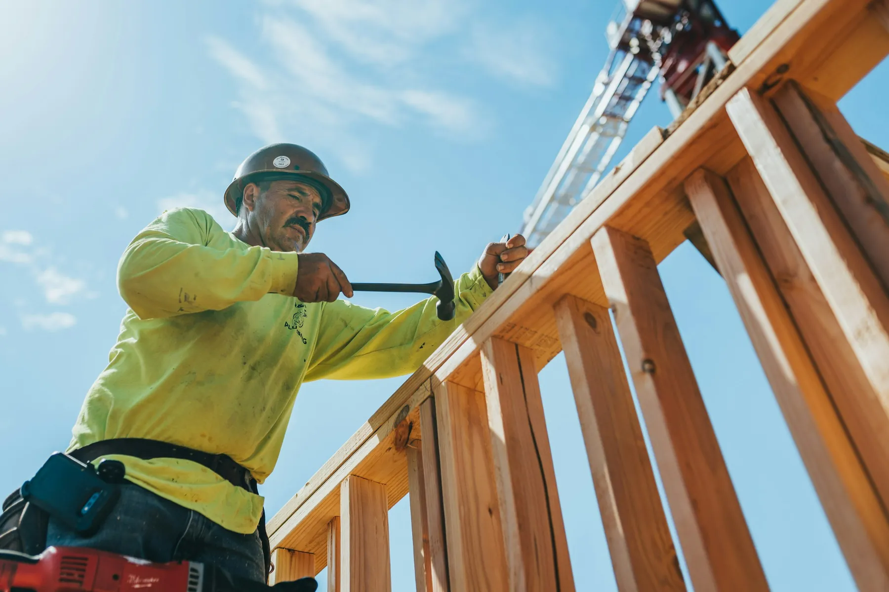 A framer in a hard hat and high-visibility shirt hammers a nail into new wall framing under a blue sky