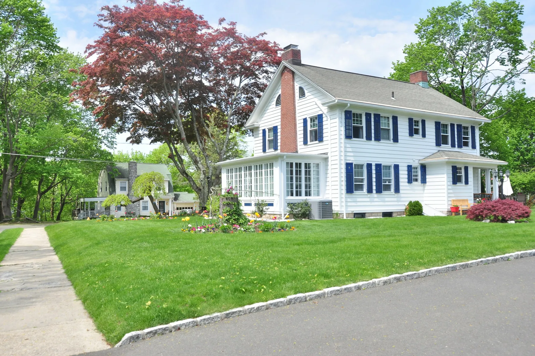 A white two-storey colonial house with navy shutters behind a sloping green lawn, framed by mature trees