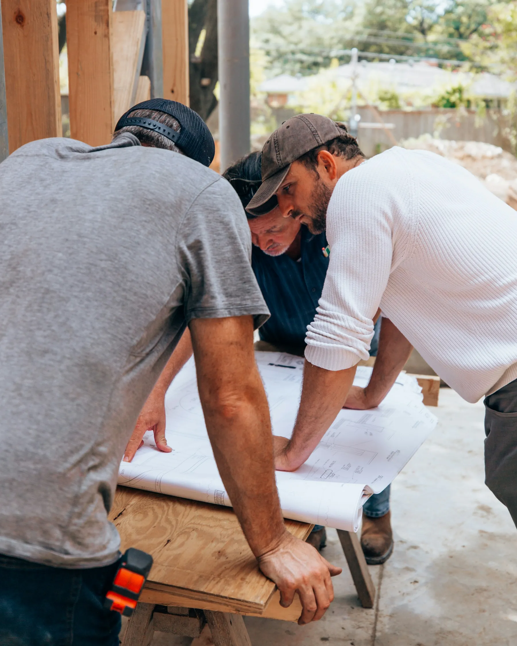 Three builders lean over a set of house plans spread on a plywood table at a job site